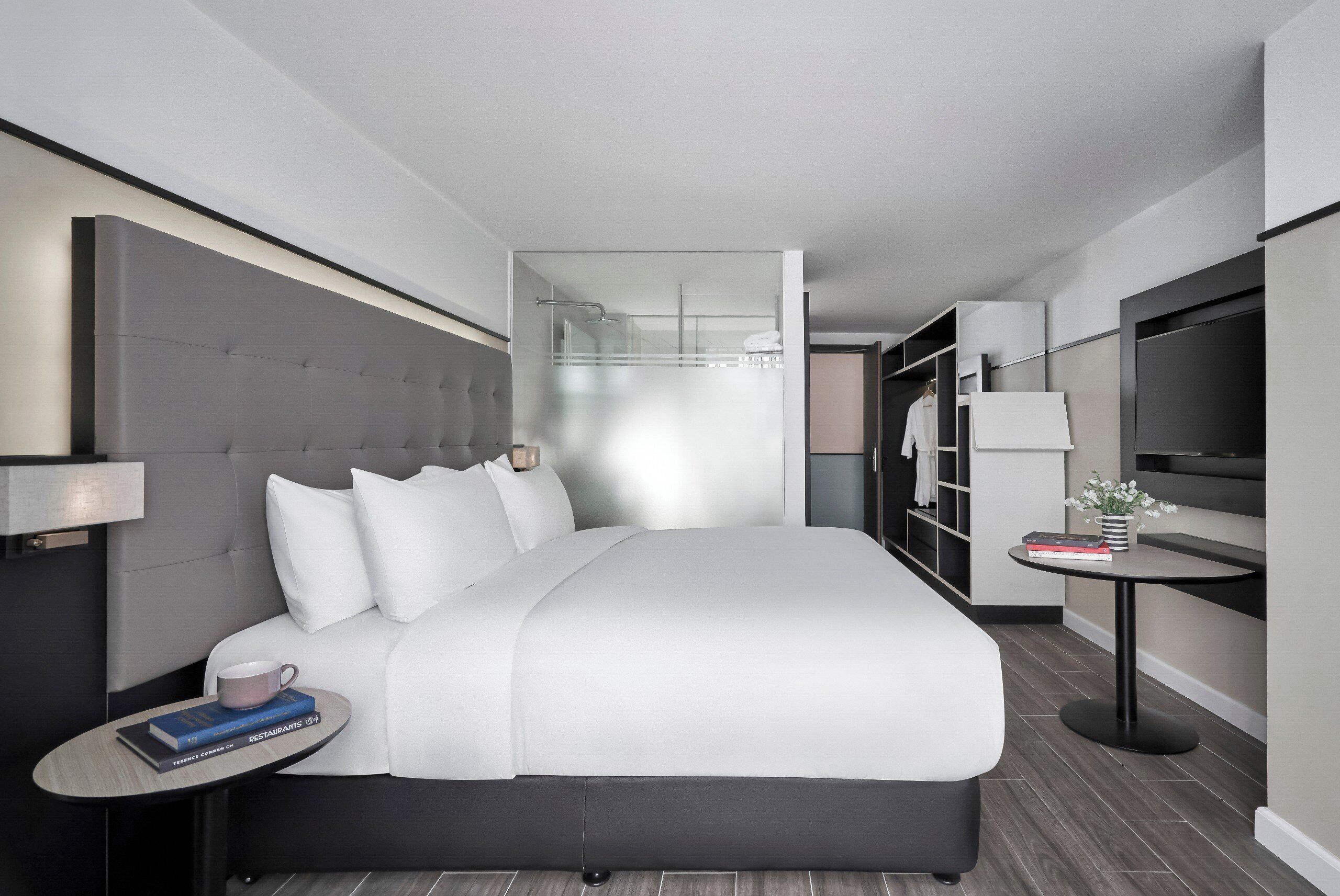 A modern hotel room with a large bed, a bedside table with a cup and books, a small round table with a vase, and a wardrobe.