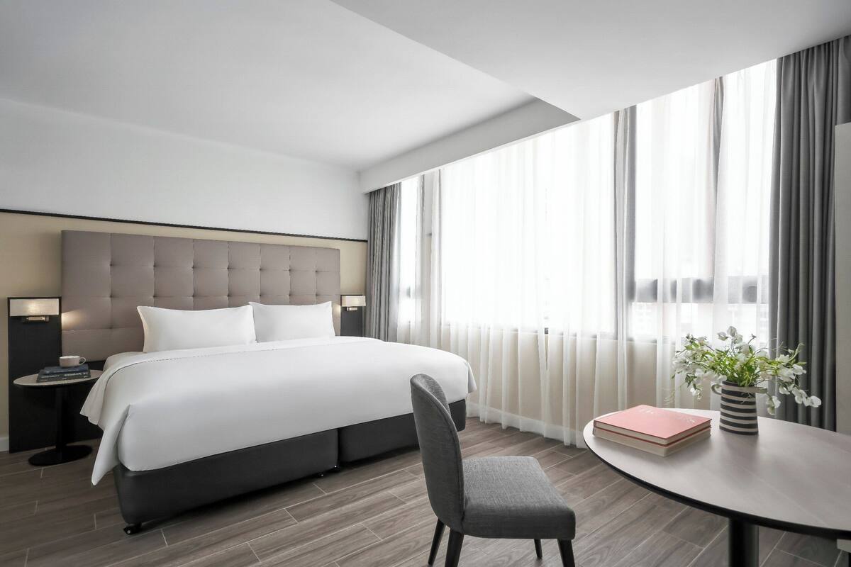 Une chambre d’hôtel moderne dotée d’un grand lit, d’une table de chevet avec une lampe, d’une chaise, d’une petite table ornée d’un vase de fleurs et offrant une vue sur la ville grâce à de grandes fenêtres.