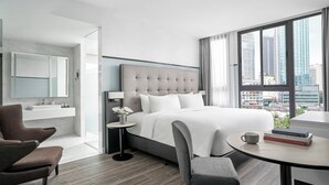 Una habitación de hotel moderna con una cama grande, un rincón de estar con una mesita de café, una mesita auxiliar pequeña y vistas a la ciudad a través de amplios ventanales.