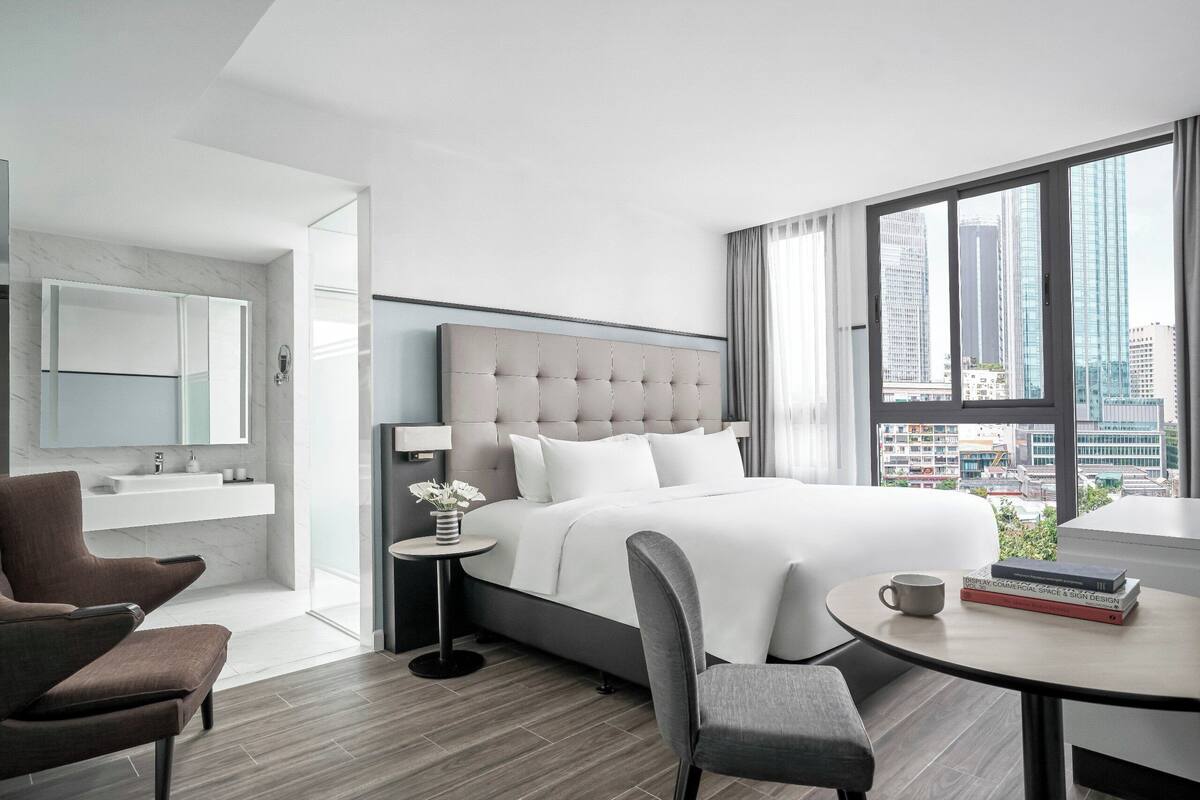 Une chambre d’hôtel moderne dotée d’un grand lit, d’un coin salon avec une table basse, une petite table d’appoint et offrant une vue sur la ville grâce à de grandes fenêtres.