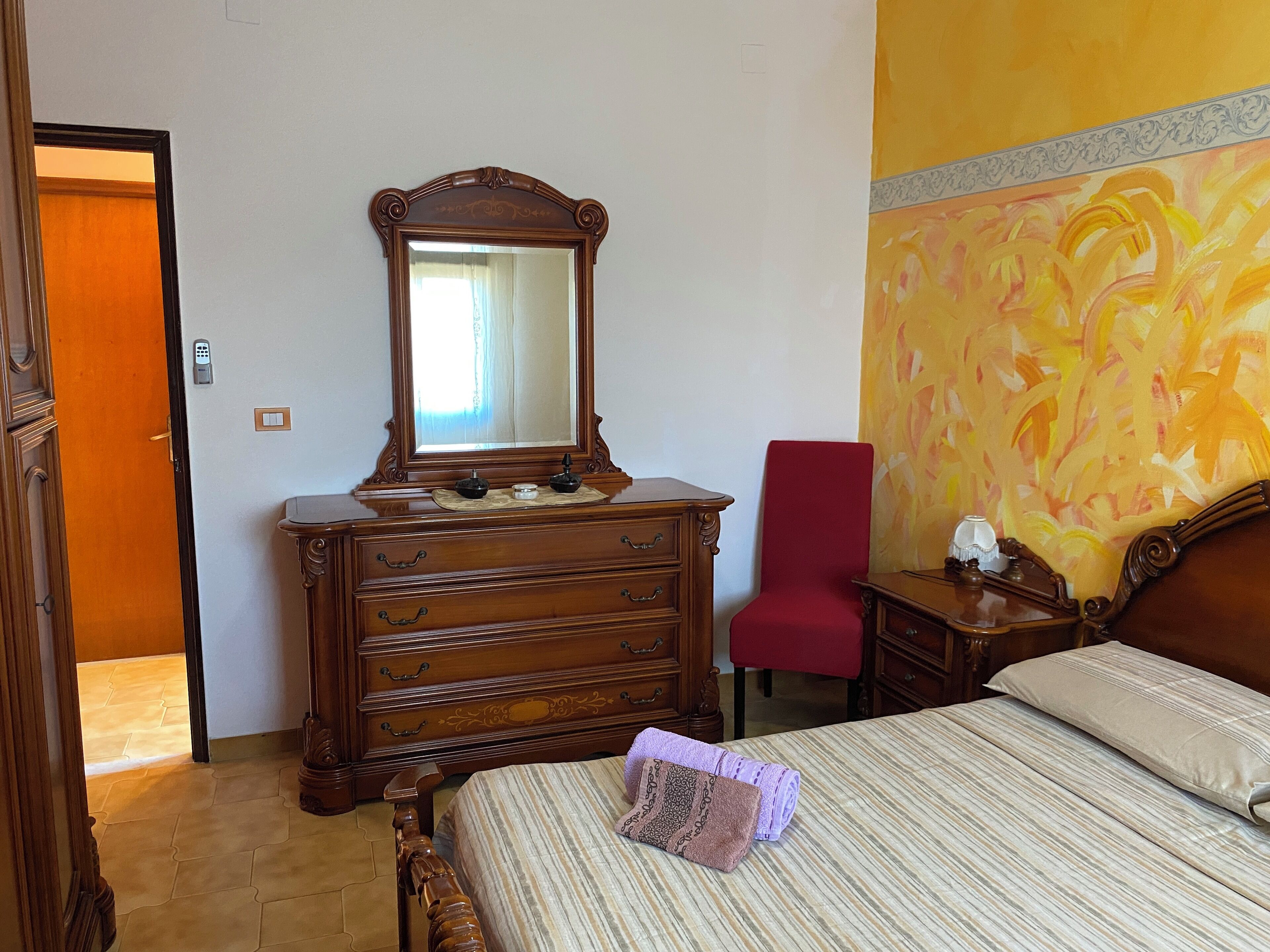 Een slaapkamer met een houten dressoir, een rode stoel, een bed met gestreept beddengoed en een grote spiegel.