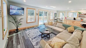 Un salon spacieux avec un grand canapé, une table basse et un téléviseur fixé au mur. De grandes fenêtres offrent une vue sur une terrasse enneigée à l’extérieur.