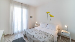 A bedroom with a bed, white curtains, a nightstand, a lamp, and a vase with yellow flowers. - Colorful beachfront apartment (Bibione)