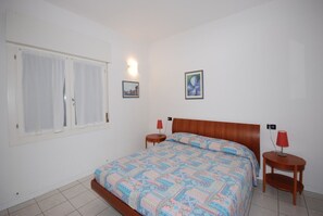 A bedroom with a wooden bed, two bedside tables with lamps, a window with curtains, and two framed pictures on the wall.