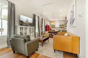 Uma sala de estar com sofá, poltrona e mesa de centro, um espaço de jantar com mesa e cadeiras, e uma cozinha com geladeira e bancada.