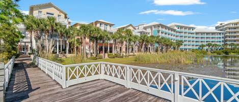 A wooden boardwalk leading to a hotel with multiple floors and balconies, surrounded by palm trees and a body of water.