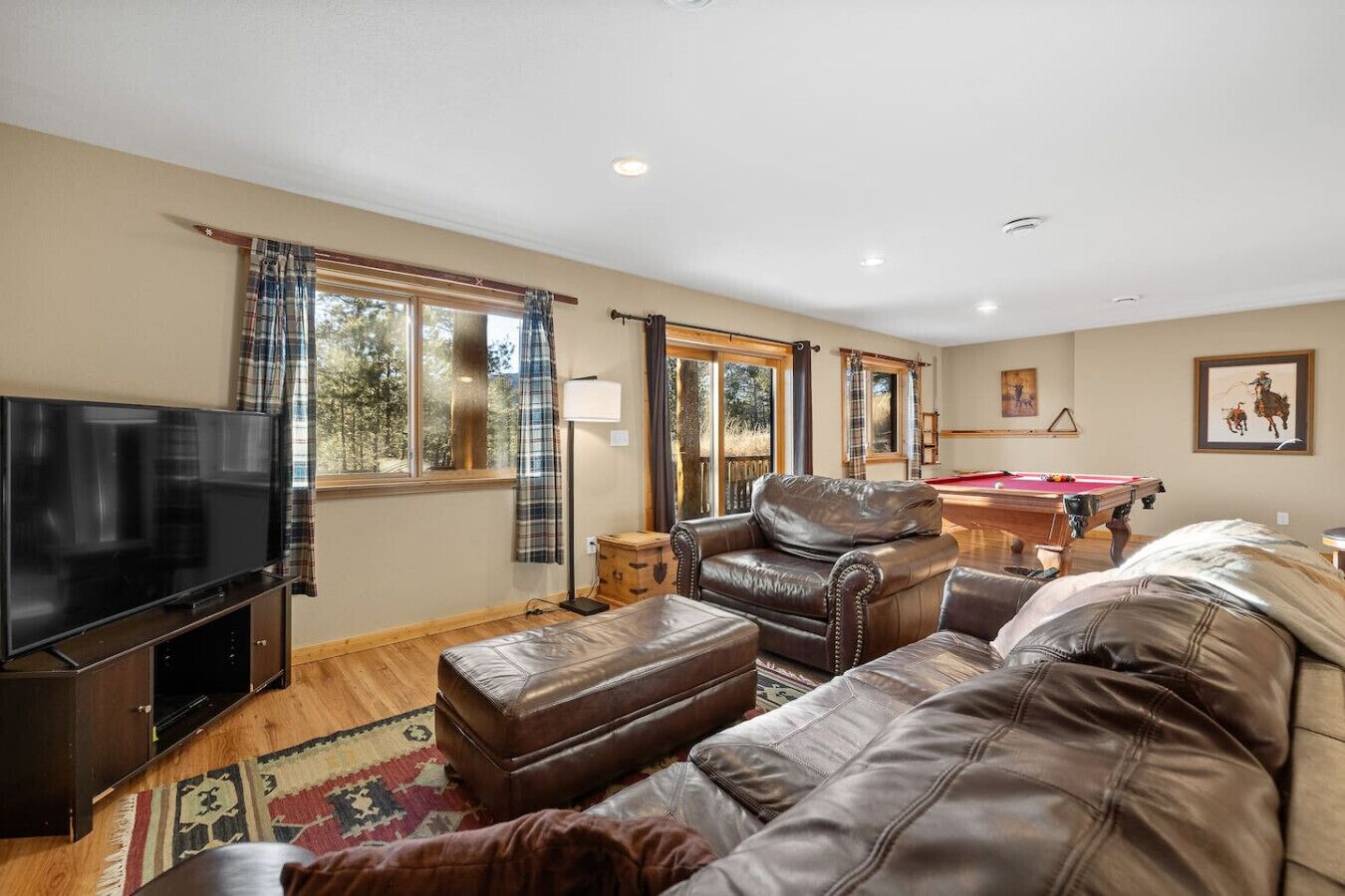 A living room with a television, leather sofas, a pool table, and large windows with curtains.