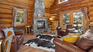 A log cabin interior with a stone fireplace, leather furniture, and large windows. - Beaver Creek Luxury Lodge (Custer)