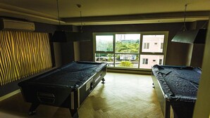 A room with two pool tables, a large window with a view of buildings and greenery, and a wooden floor. - GSR - Grande Caribbean (Pattaya)