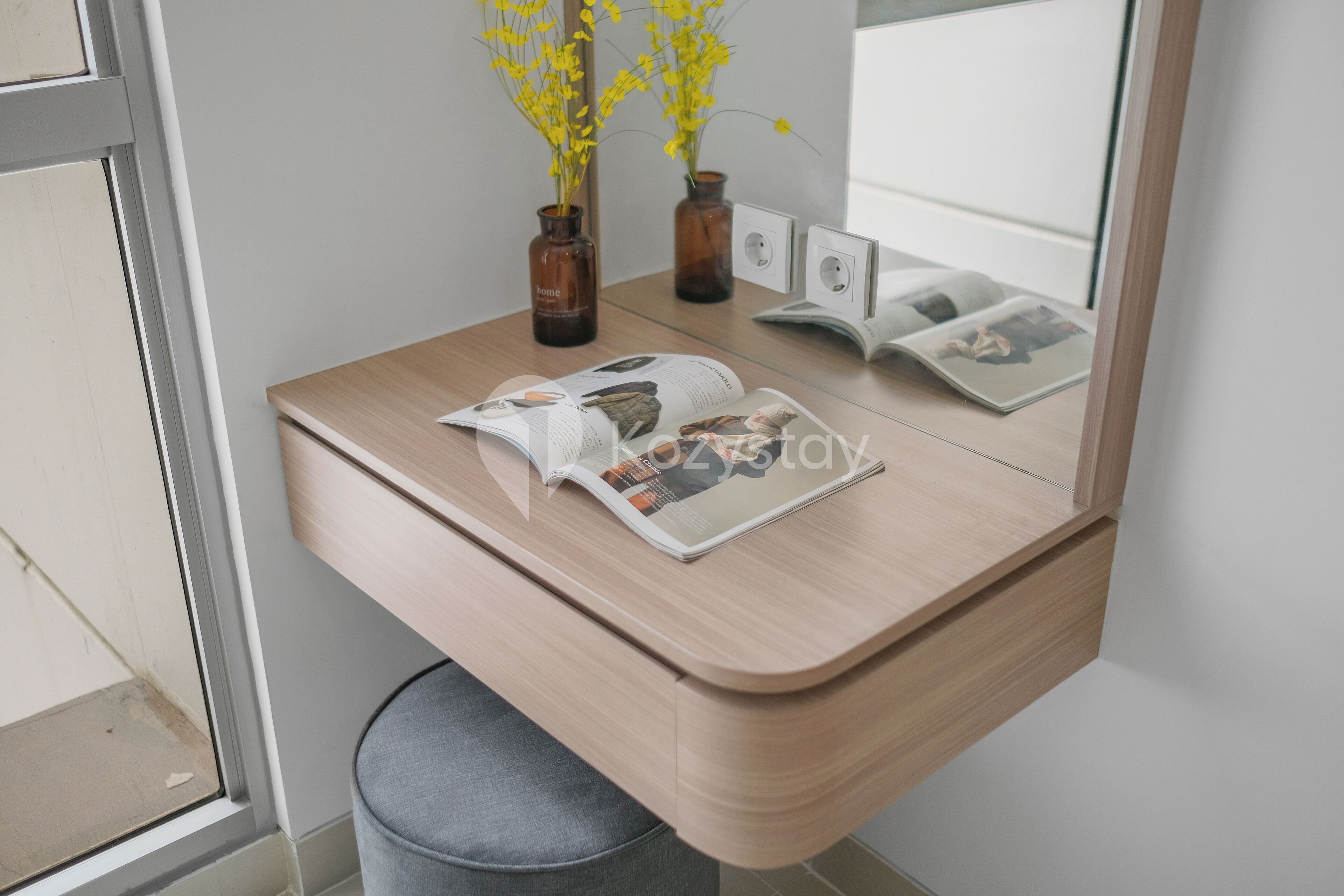 Un escritorio moderno con una revista, dos jarrones con flores amarillas y un cuadro enmarcado sobre una superficie de madera clara.