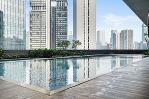 Una piscina en la azotea con cubierta de madera, rodeada de vegetación y vistas a un paisaje urbano con edificios altos.