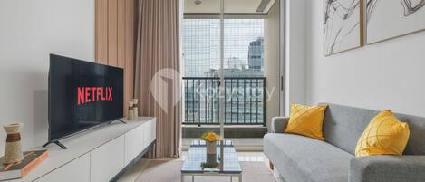 A modern living room with a flat-screen TV displaying 'NETFLIX', a grey sofa with yellow cushions, a glass coffee table, and a city view through large windows.