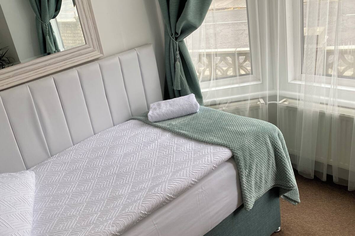 A bed with a white mattress and green bedding, a mirror with a white frame, and a window with sheer curtains.