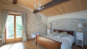 Un dormitorio con pared de piedra, vigas de madera en el techo, una cama grande con almohadones florales, una mesita de noche con lámpara y una ventana con cortinas.