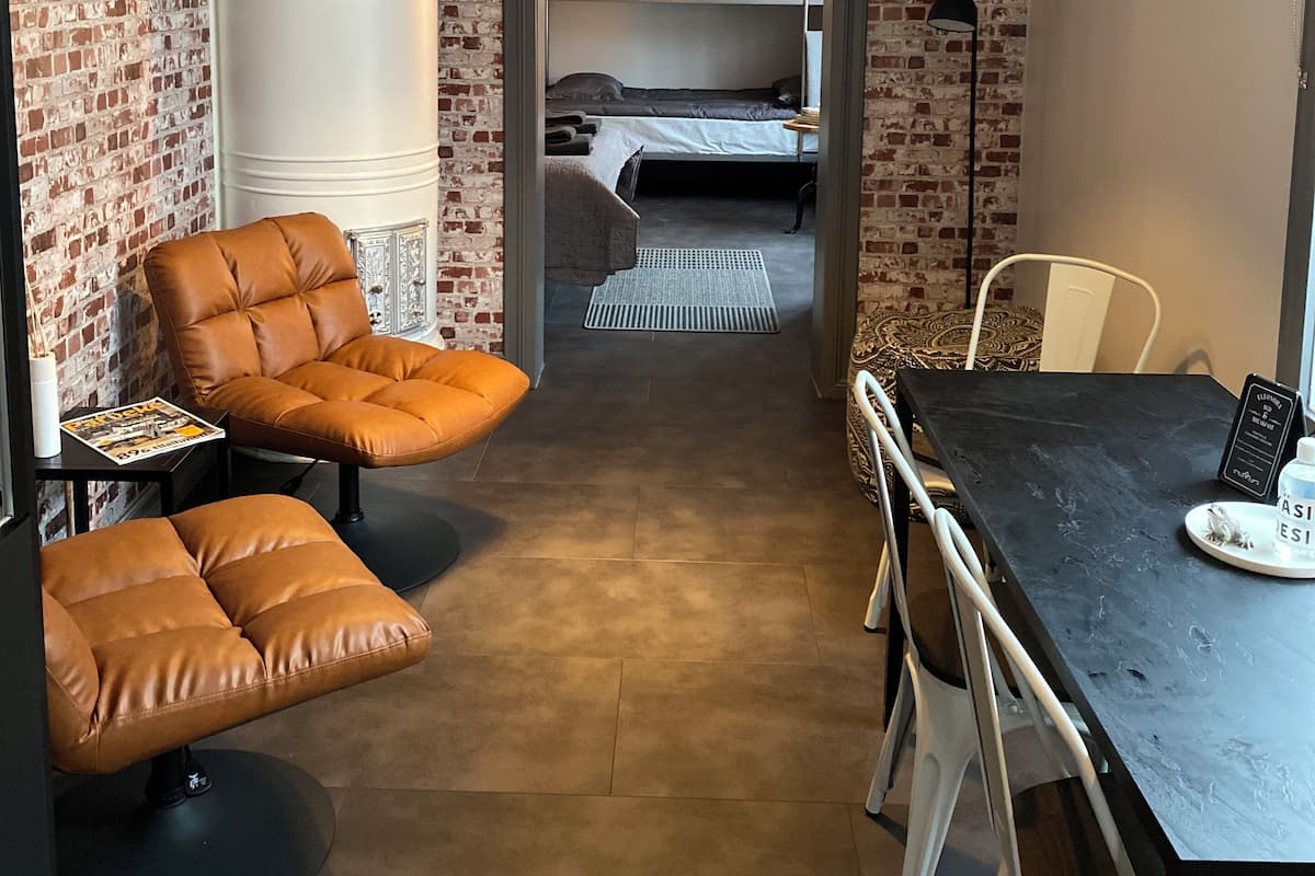 A compact room with a brick wall, a bunk bed, a dining table with chairs, a leather armchair, and a pendant light.