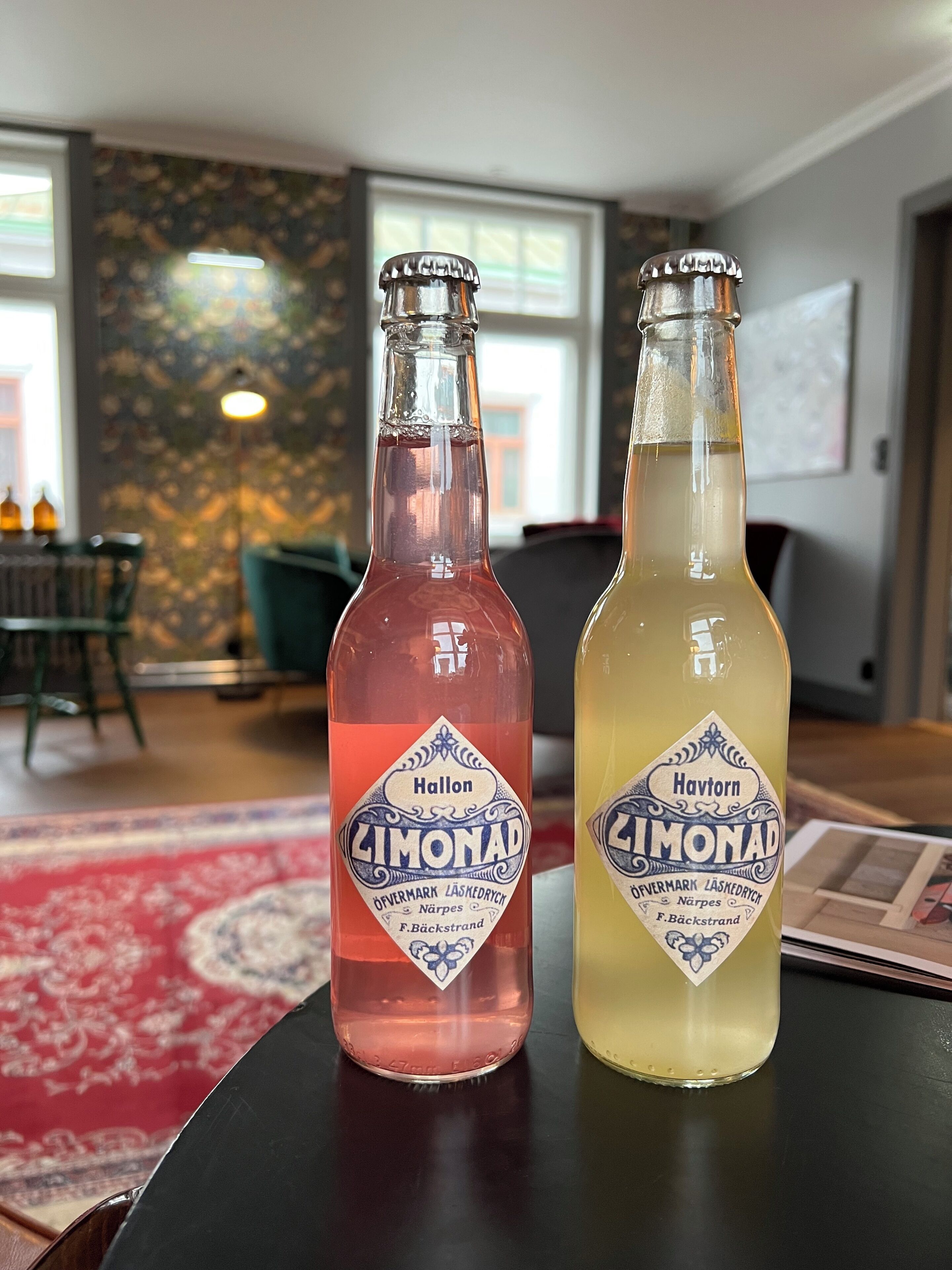Two bottles of Hallon Limonad on a table in a room with a patterned wall and a window.