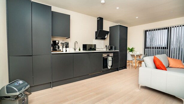 A modern kitchen with dark cabinets, a white countertop, and a built-in oven. There is a dining area with a white sofa and a small table.