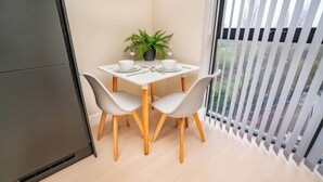A small dining area with a table set for two, a potted plant, and a view of the outdoors through a window with blinds.