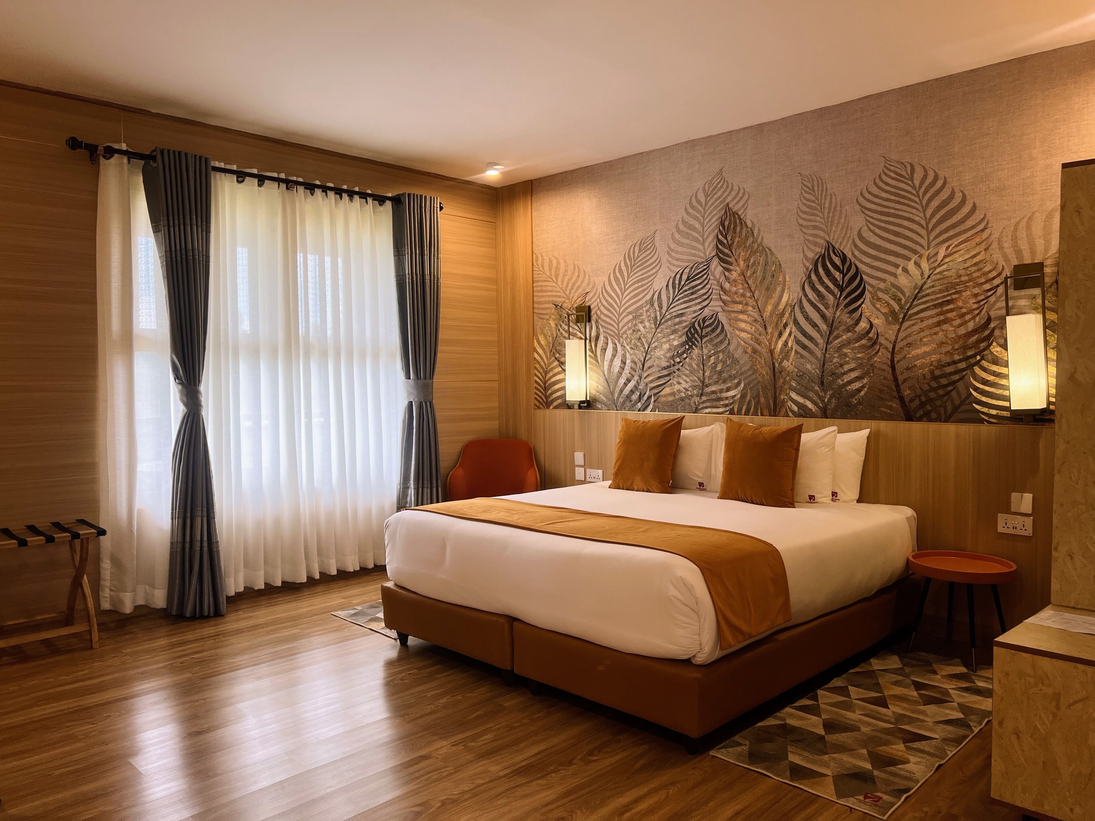 A hotel room with a large bed, wooden flooring, a wall-mounted lamp, and a leaf-patterned mural.