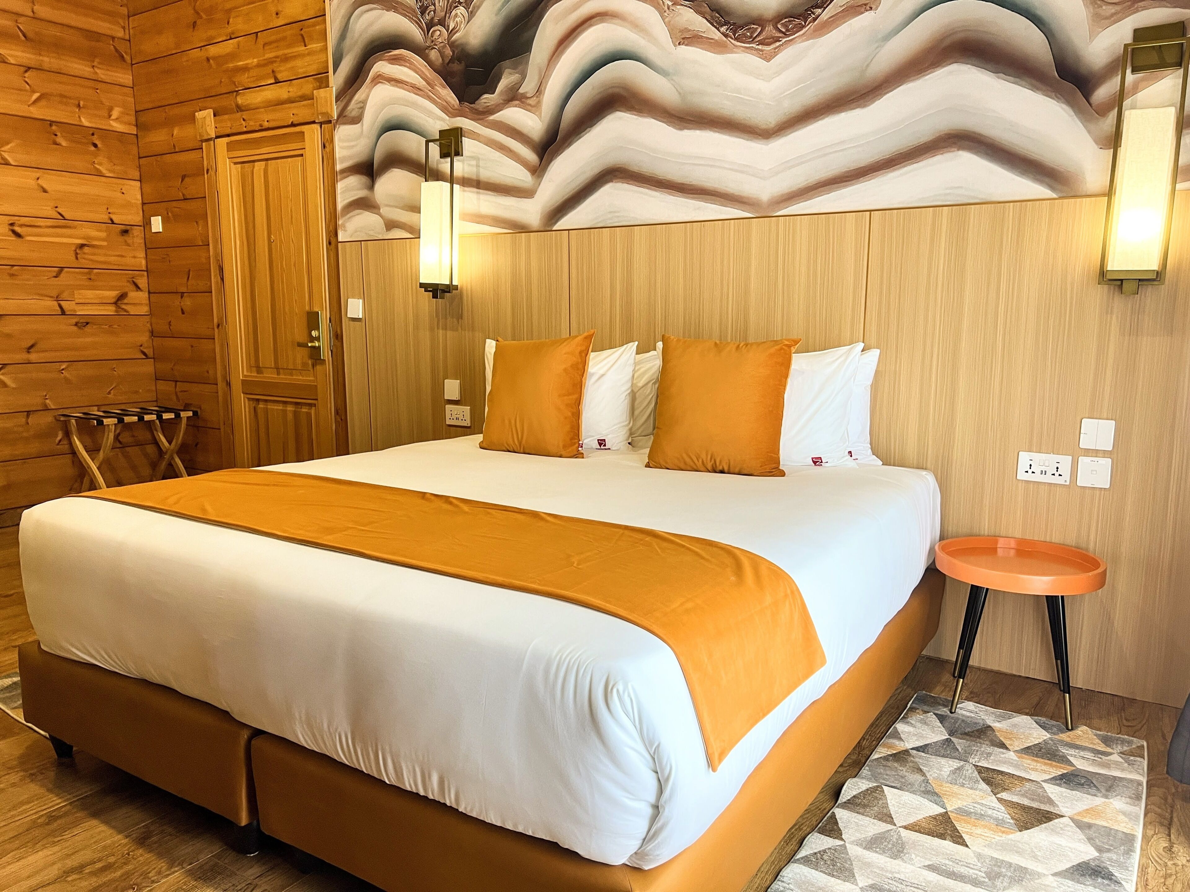 A hotel room with a large bed, wooden walls, a decorative ceiling mural, a small round table, and a patterned rug.