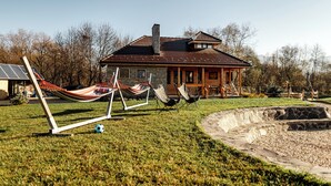 Une maison avec une façade en pierre, un porche en bois et une cheminée en pierre. On trouve deux hamacs et deux chaises pliantes dans la cour.