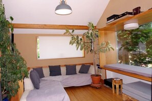 Ein gemütliches Zimmer mit Holzboden, einem großen Fenster, einer Couch mit Kissen, einer Zimmerpflanze und einem kleinen Hocker.