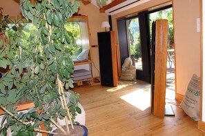 Une piĂšce avec une grande plante en pot, un lit donnant sur une vue agrĂ©able, un haut-parleur noir et une porte-fenĂȘtre coulissante qui mĂšne Ă lâextĂ©rieur.