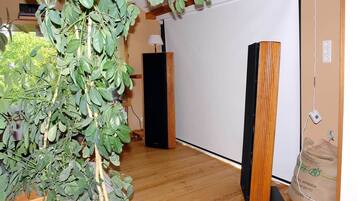 Una habitación con suelo de madera, una planta grande, un altavoz negro y una pizarra.