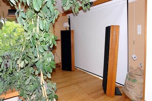 Ein Zimmer mit Holzboden, einer großen Pflanze, einem schwarzen Lautsprecher und einer Whiteboard.