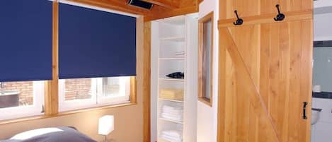A bedroom with a bed, a window with a blind, a wooden wardrobe, and a white shelf unit.