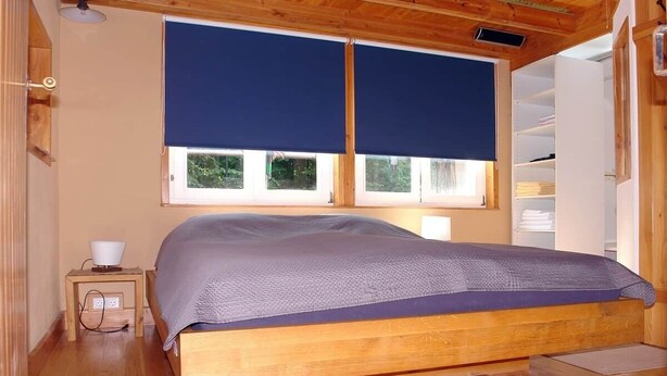 A wooden bed with a blue bedspread, a bedside table with a lamp, and a window with blinds.
