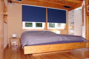 Uma cama de madeira com colcha azul, uma mesa de cabeceira com abajur e uma janela com persianas.