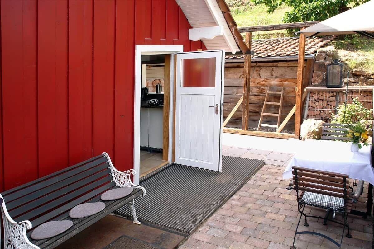 A red building with a white door, a wooden bench, a table with a white tablecloth, and a small garden area.
