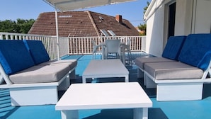 A covered outdoor seating area with two lounge chairs and a small table, a white railing, and a view of a house with a tiled roof.
