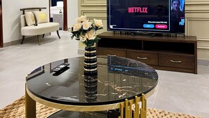A living room with a flat-screen TV displaying Netflix, a sofa, a coffee table with a vase of flowers, and a decorative mirror.