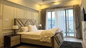 A modern bedroom with a large bed, bedside lamps, a nightstand, and a view of the city through large windows.