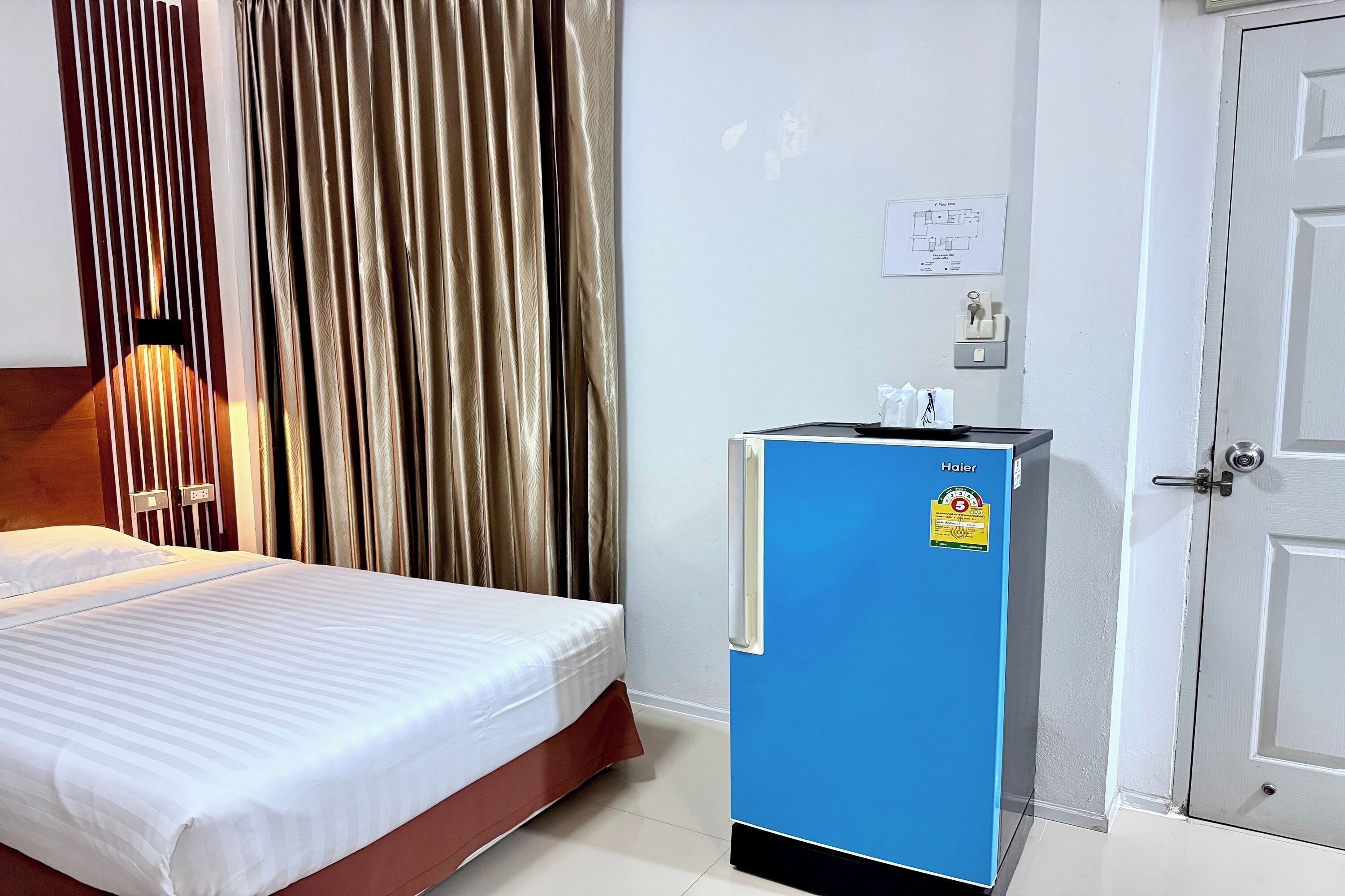 Habitación de hotel con cama, cortinas, minibar azul, una puerta y un interruptor de luz en la pared.