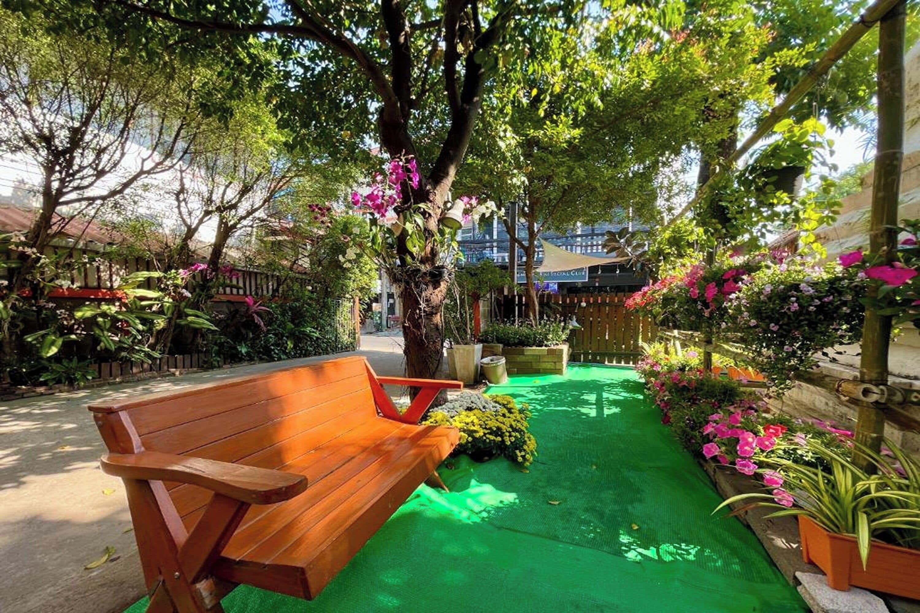 A wooden bench in a garden with green artificial turf, surrounded by flowering plants and trees.