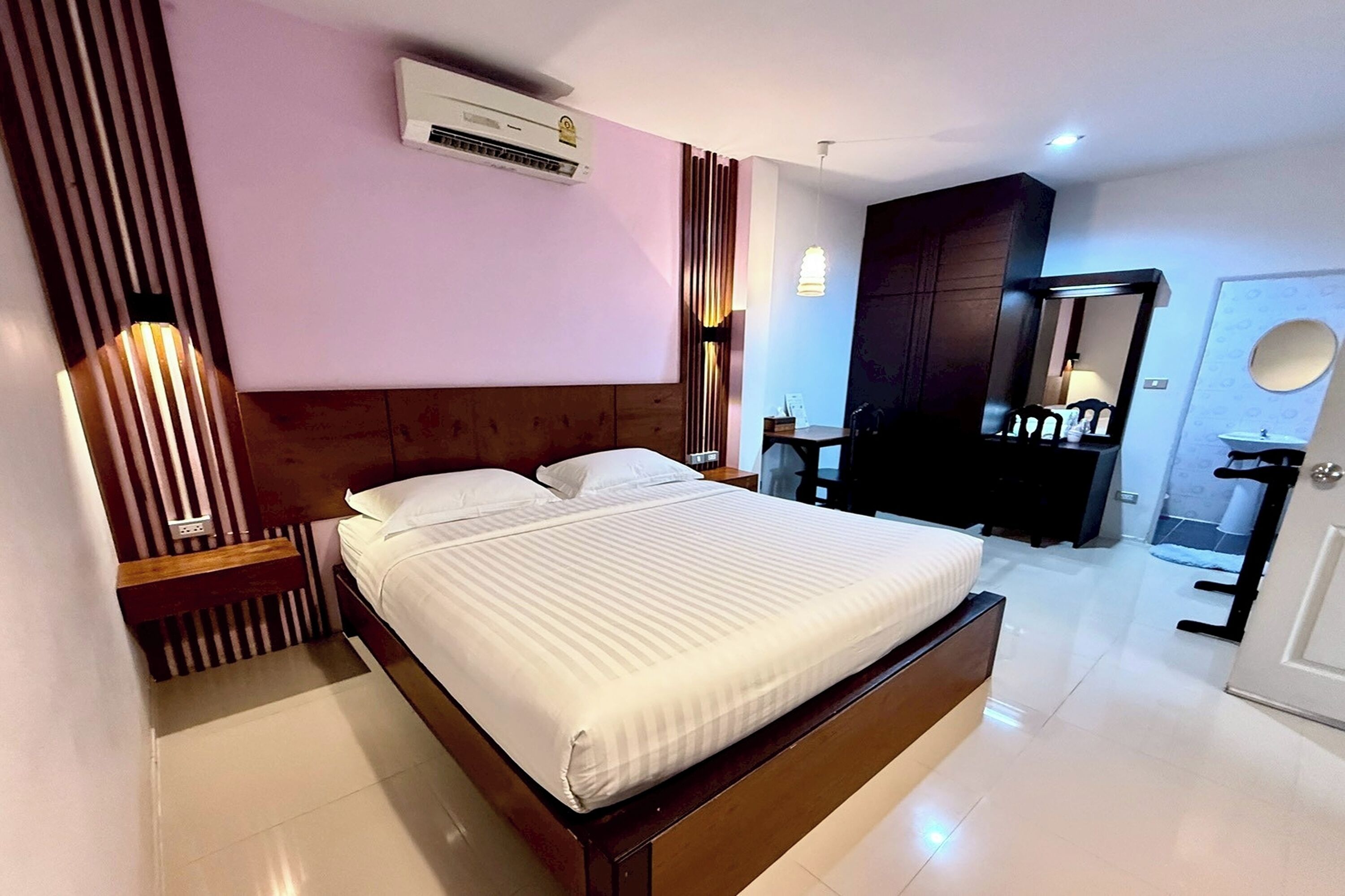 A modern hotel room with a large bed, a wooden headboard, a bedside table, a wardrobe, a mirror, and a bathroom with a sink and toilet.
