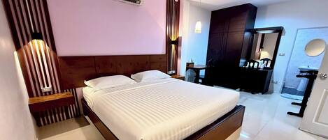 A modern hotel room with a large bed, a wooden headboard, a bedside table, a wardrobe, a mirror, and a bathroom with a sink and toilet.