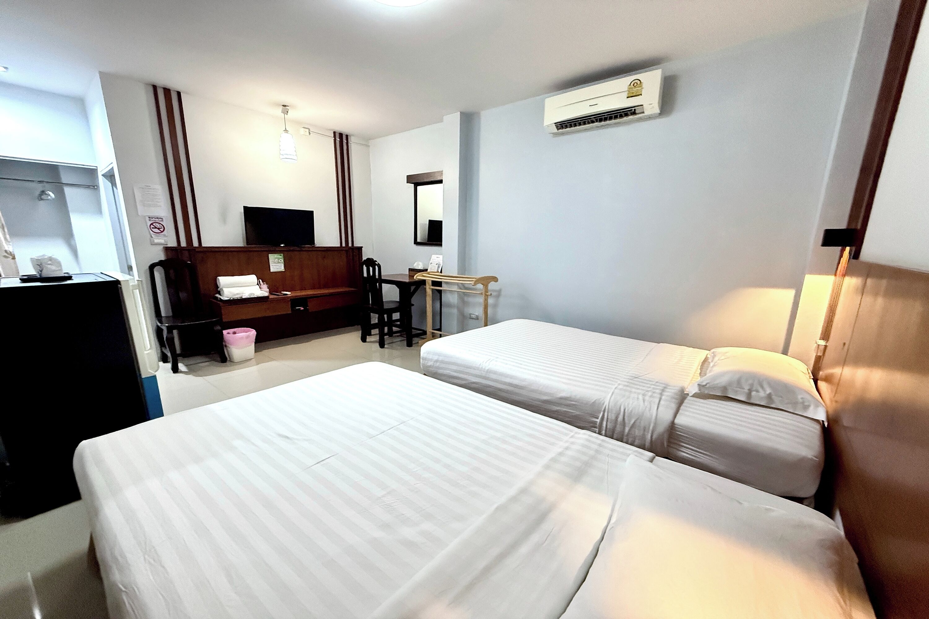 A hotel room with two beds, a desk, a chair, a wardrobe, a TV, and an air conditioning unit.