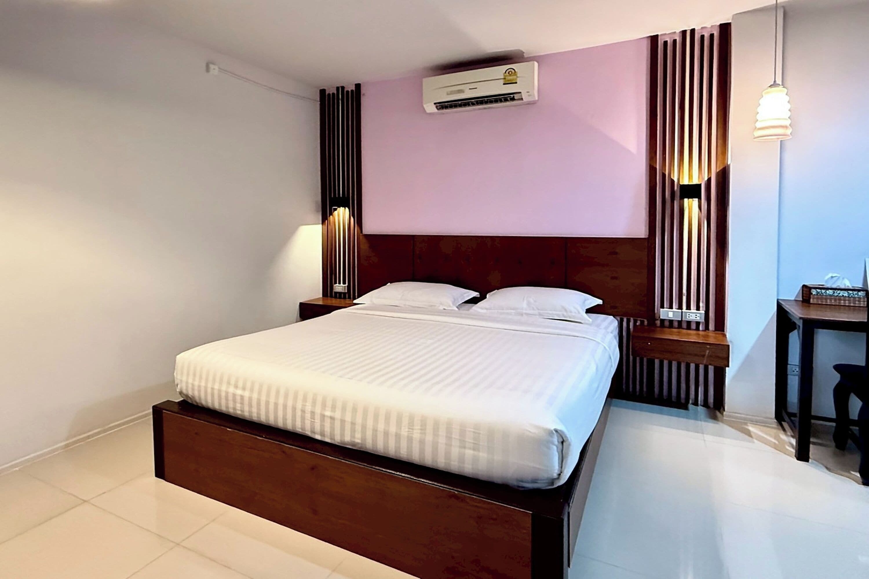 A neatly made bed in a hotel room with a wooden headboard, a wall-mounted air conditioner, and a desk with a chair.