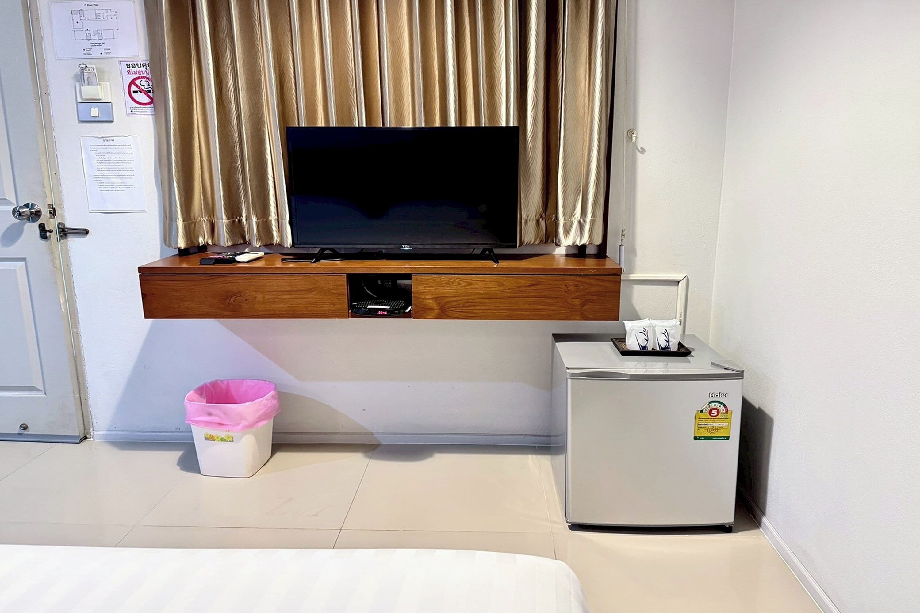 A hotel room with a television mounted on a wooden shelf above a desk, a small refrigerator, and a trash bin.