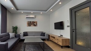 Ein modernes Wohnzimmer mit einem grauen Sofa, einem kleineren grauen Sessel, einem Couchtisch, einer Holz-TV-Bank und einem großen Fenster mit Jalousien.