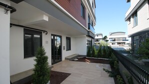 A modern building with a clean, white exterior, a tiled patio area, and a small garden with a coniferous tree. - Professionally Maintained Spacious New Suites 2-6 (Antalya)