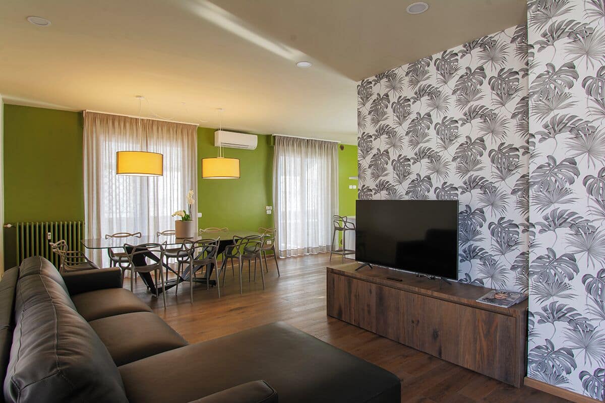 A living room with a sofa, a TV on a wooden stand, a dining area with chairs and a table, and a wall with tropical wallpaper.
