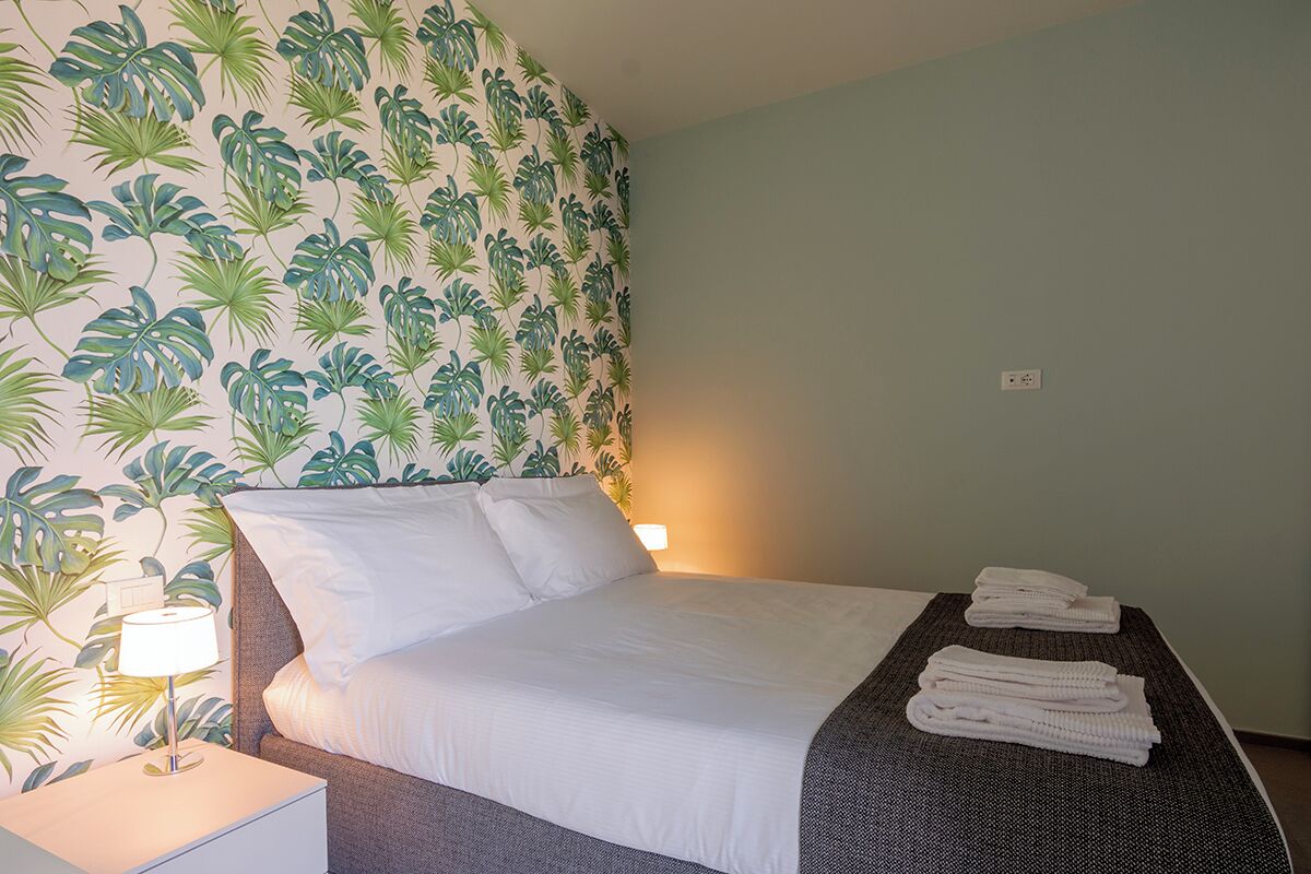 A neatly made bed with white linens, a bedside lamp, and a stack of towels on a dark headboard.