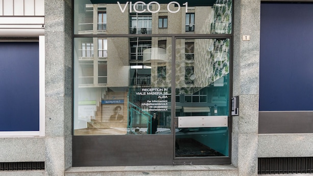 Una entrada de edificio con área de recepción, una escalera y fachada de vidrio que refleja el interior.