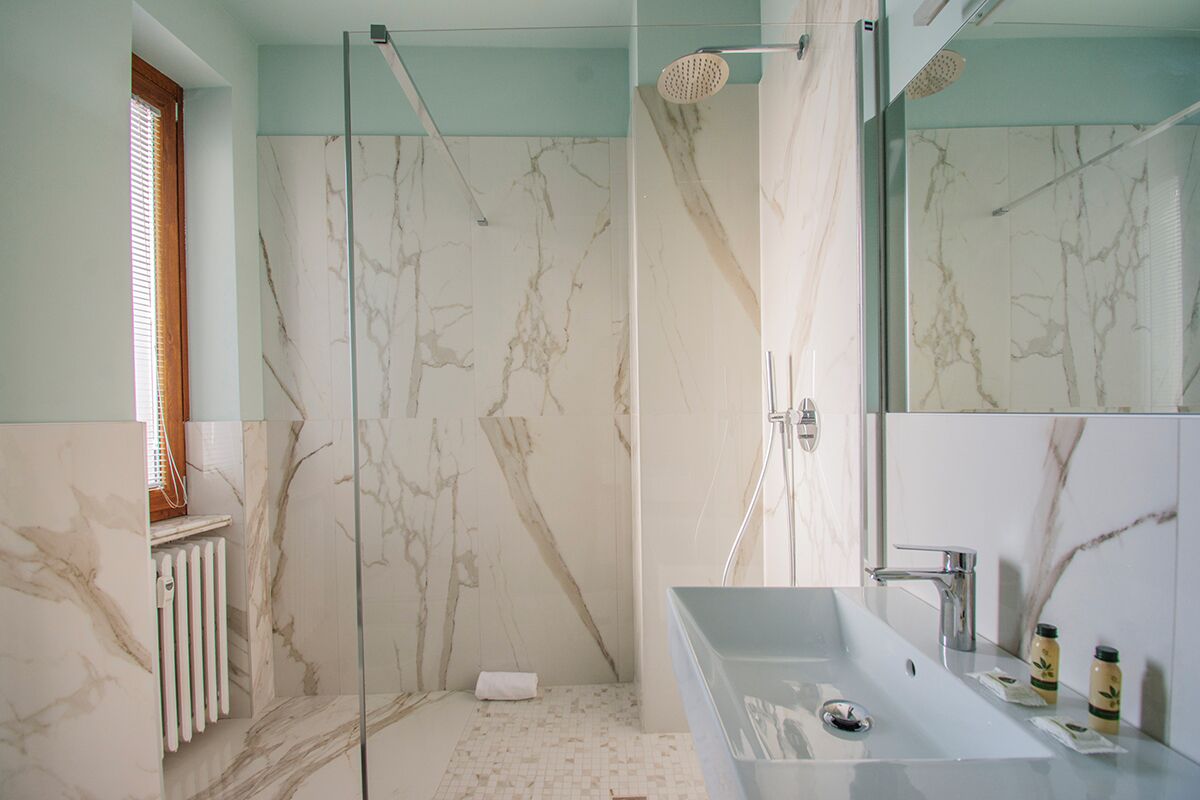 Une salle de bain moderne dotée d’une douche à parois de verre, d’un mur de douche recouvert de carreaux de marbre, d’un grand miroir et d’un lavabo avec des articles de toilette.
