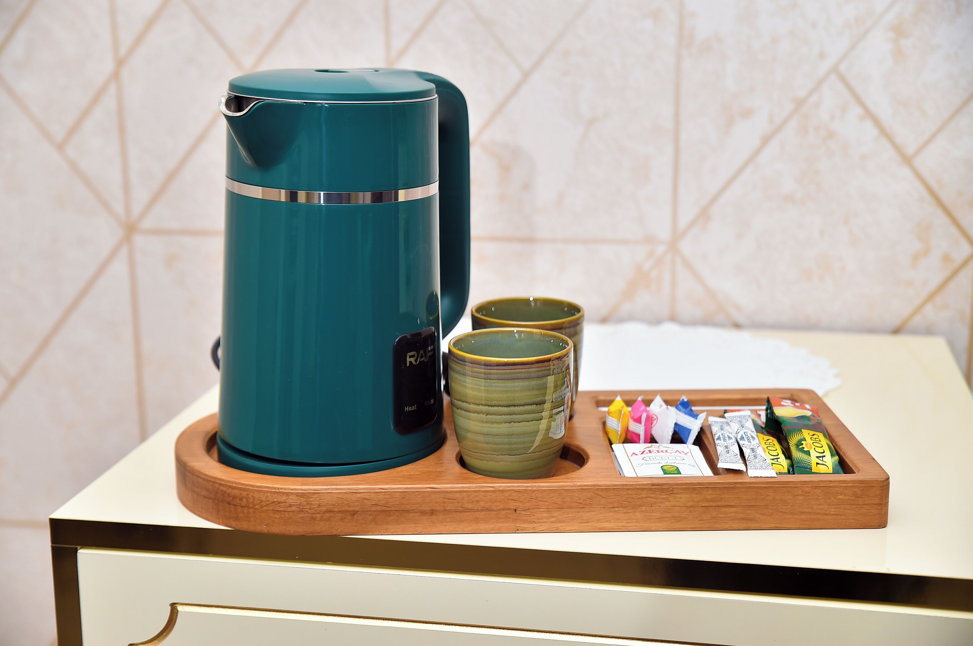 Una tetera eléctrica color turquesa, dos tazas de cerámica verde y una bandeja con varios objetos, incluyendo una caja con la etiqueta "JACOBS" sobre una bandeja de madera.
